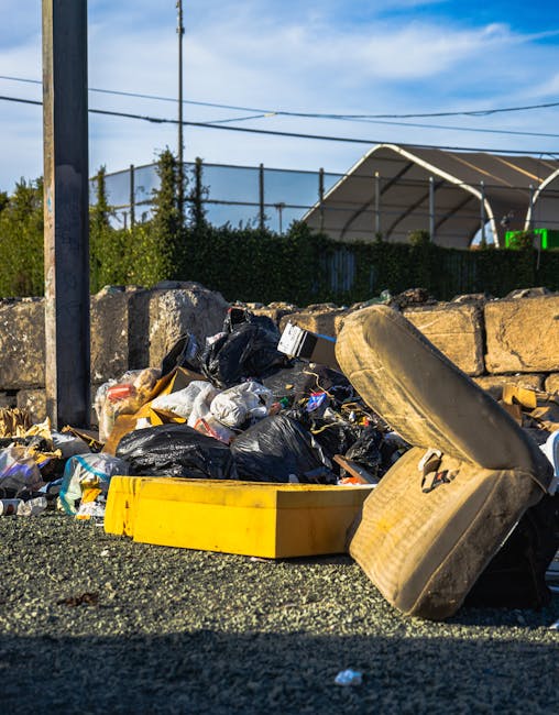 A pile of mixed waste and rubbish items situated on the side of a paved area next to a low stone wall. The waste includes multiple black plastic bin bags, some torn open revealing contents, and a yell